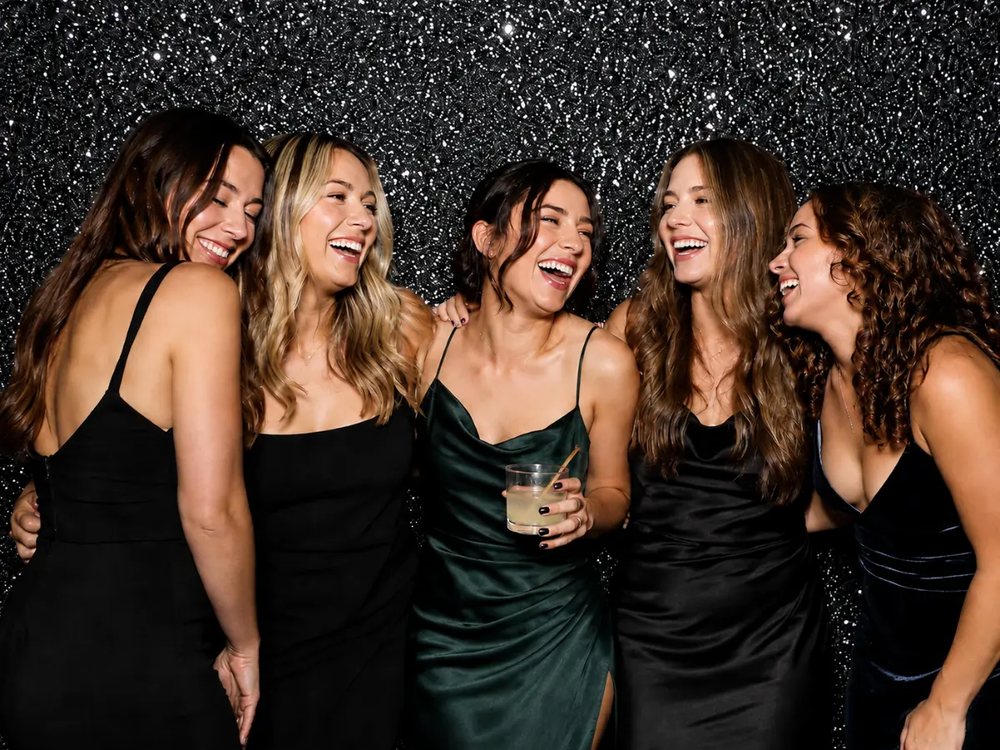 Five women in black and emerald dresses laughing at a black sparkle backdrop photo booth at a birthday party in Suffolk