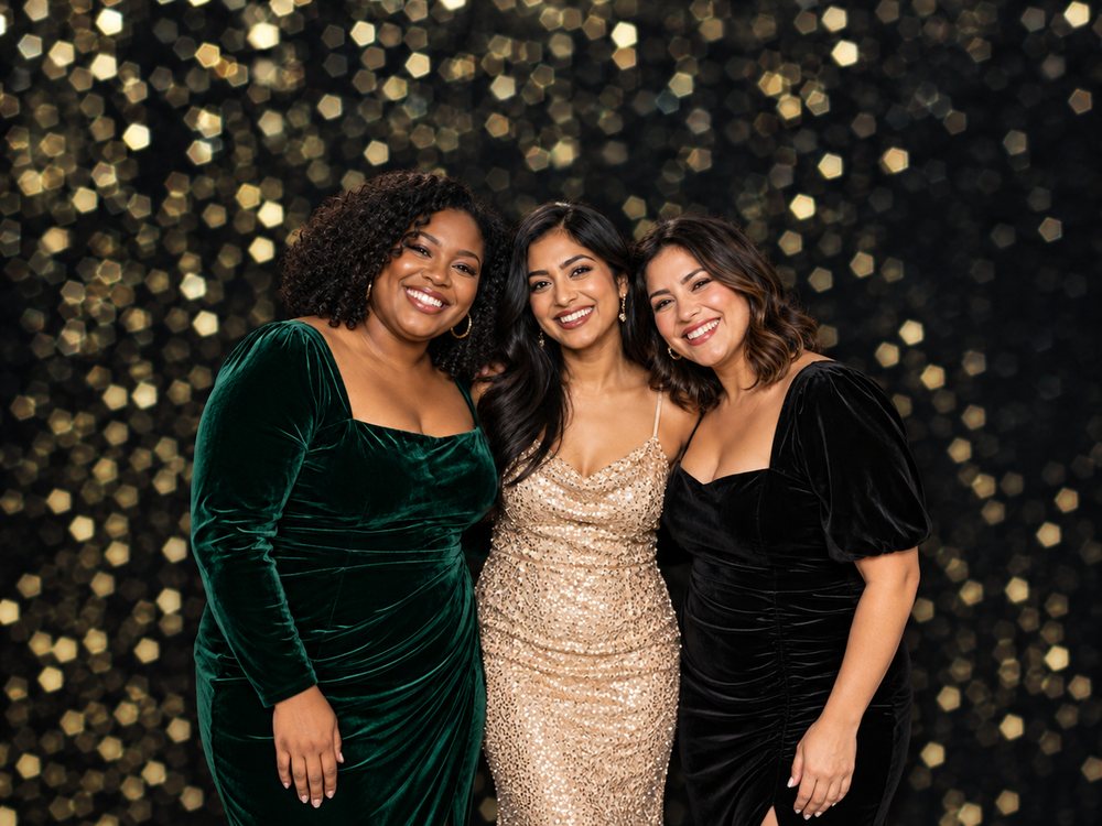 Three friends in velvet and sequin dresses at a gold bokeh holiday backdrop photo booth at a corporate holiday party in Norfolk
