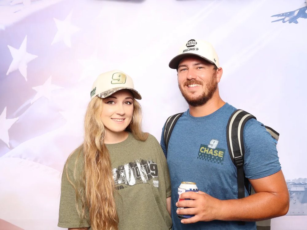 A couple in racing caps smiling at a custom flag and aircraft carrier backdrop photo booth at the Newport News Shipbuilding Cook Out 400 at Richmond Raceway in Richmond