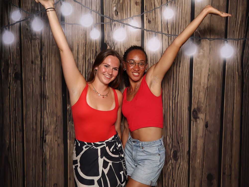 Two friends with arms up under string lights at a dark wood backdrop Fourth of July party photo booth in Virginia Beach