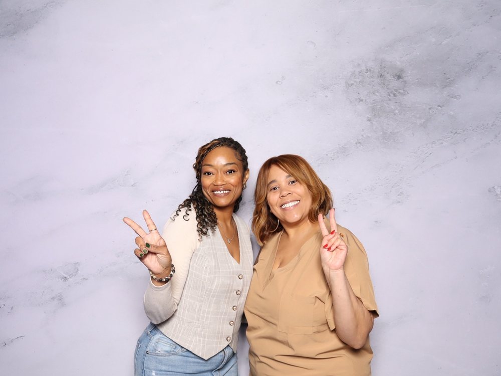 Two friends smiling and throwing peace signs at the white marble backdrop photo booth at a bridal expo in Virginia Beach