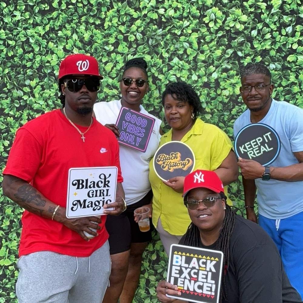 Friends in red caps holding Keep It Real, Black Girl Magic, and Black Excellence signs at an Easter family barbecue photo booth in Colerain, North Carolina