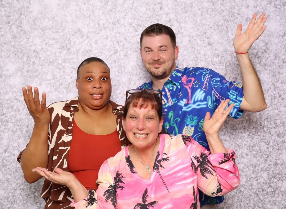 Three friends in tropical shirts waving at a pool party photo booth in Newport News