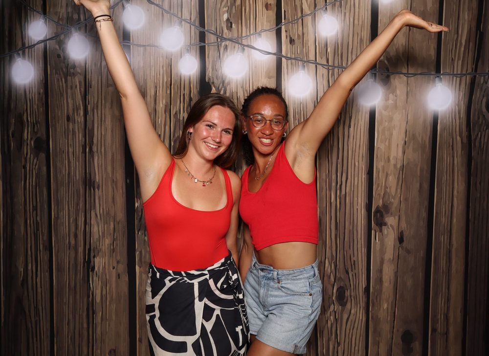 Two friends with arms up under string lights at a Fourth of July party photo booth in Virginia Beach