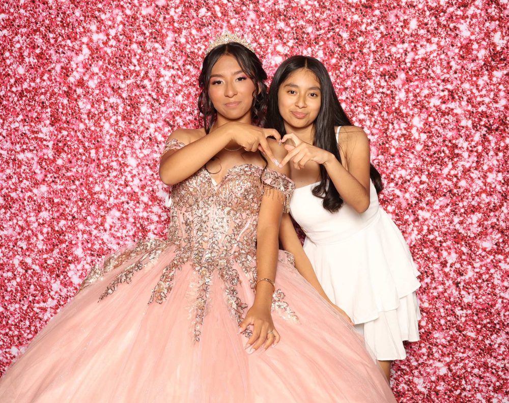 A quinceañera and a friend making heart hands at a flower-wall photo booth in Perth Amboy
