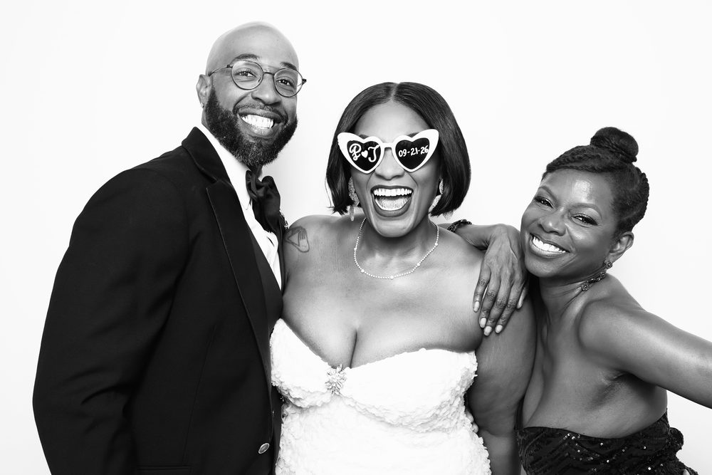 A bride in heart sunglasses with her groom and a friend at a Hampton wedding photo booth