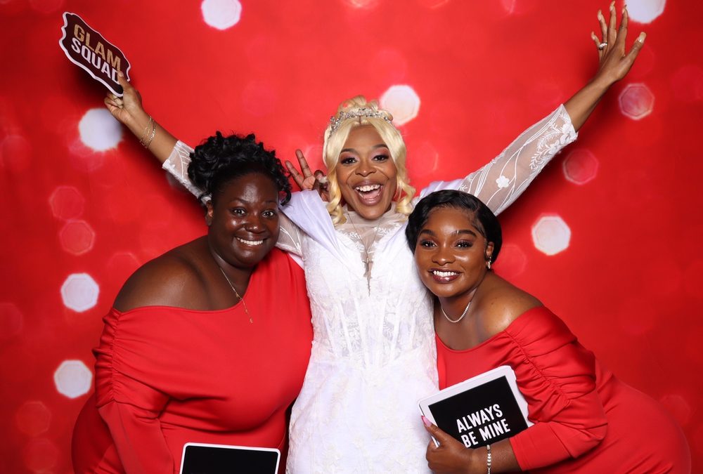 A bride with her arms up between two bridesmaids at a wedding photo booth in Suffolk