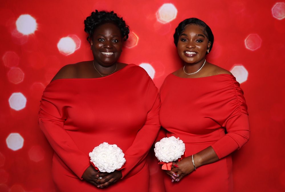 Two bridesmaids holding bouquets at a red-backdrop wedding photo booth in Suffolk