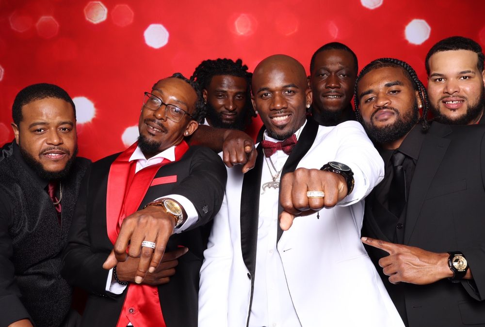A groom and groomsmen showing off rings at a red-backdrop wedding photo booth in Suffolk