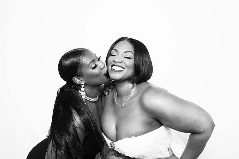 A bride and her friend sharing a cheek kiss at a black-and-white wedding photo booth in Hampton