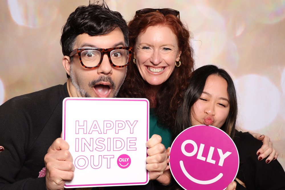 Three coworkers holding Happy Inside Out and OLLY signs at a corporate event photo booth in San Francisco