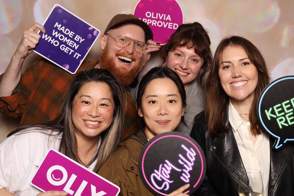 Five coworkers with Made by Women Who Get It, Olivia Approved, and Stay Wild signs at an OLLY corporate event photo booth in San Francisco