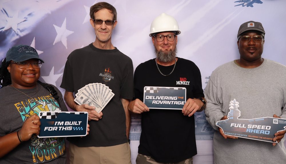 Four race fans holding Full Speed Ahead and Delivering the Advantage signs at the Newport News Shipbuilding photo booth at Richmond Raceway in Richmond